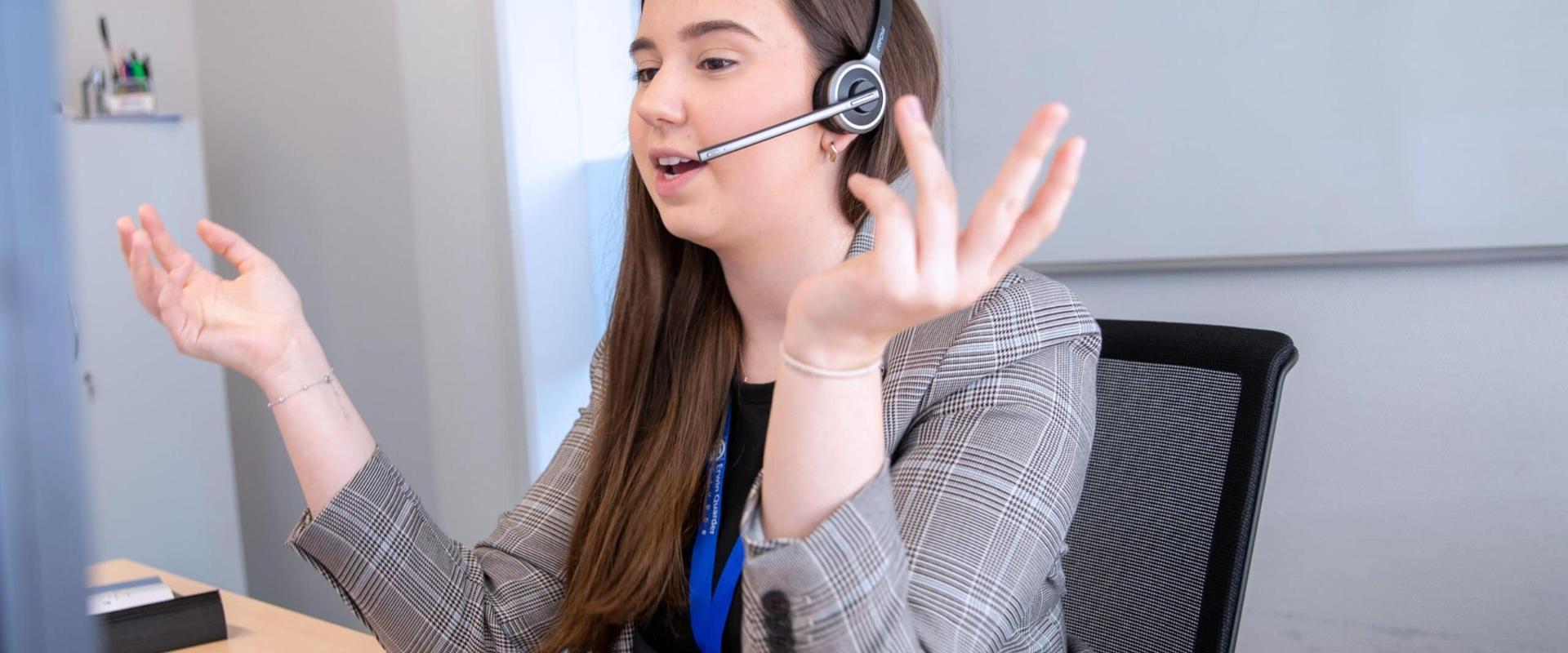 Frau mit Headset, gestikulierend, in kariertem Blazer, vor einem Schreibtisch sitzend.