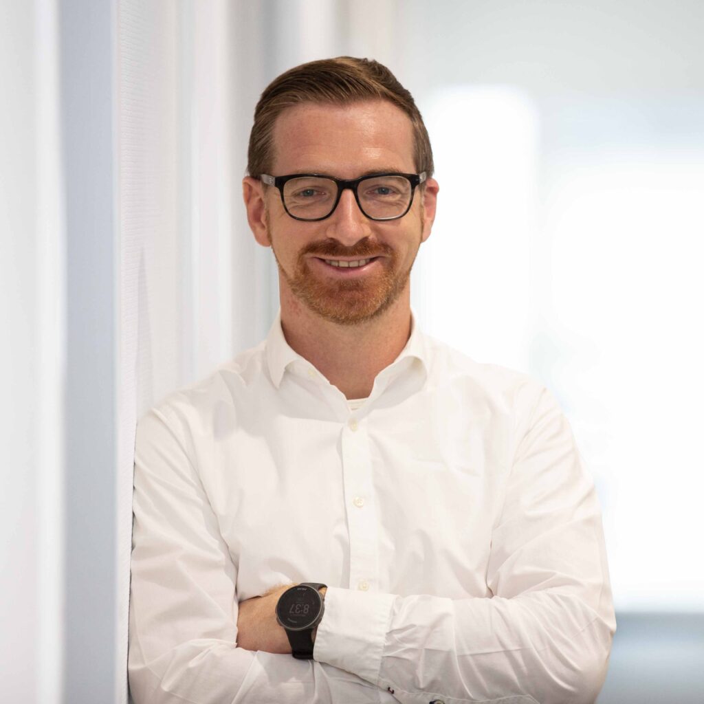 Mann mit Brille, lächelt freundlich, trägt weißes Hemd, schwarzes Armband, steht vor weißem Hintergrund.