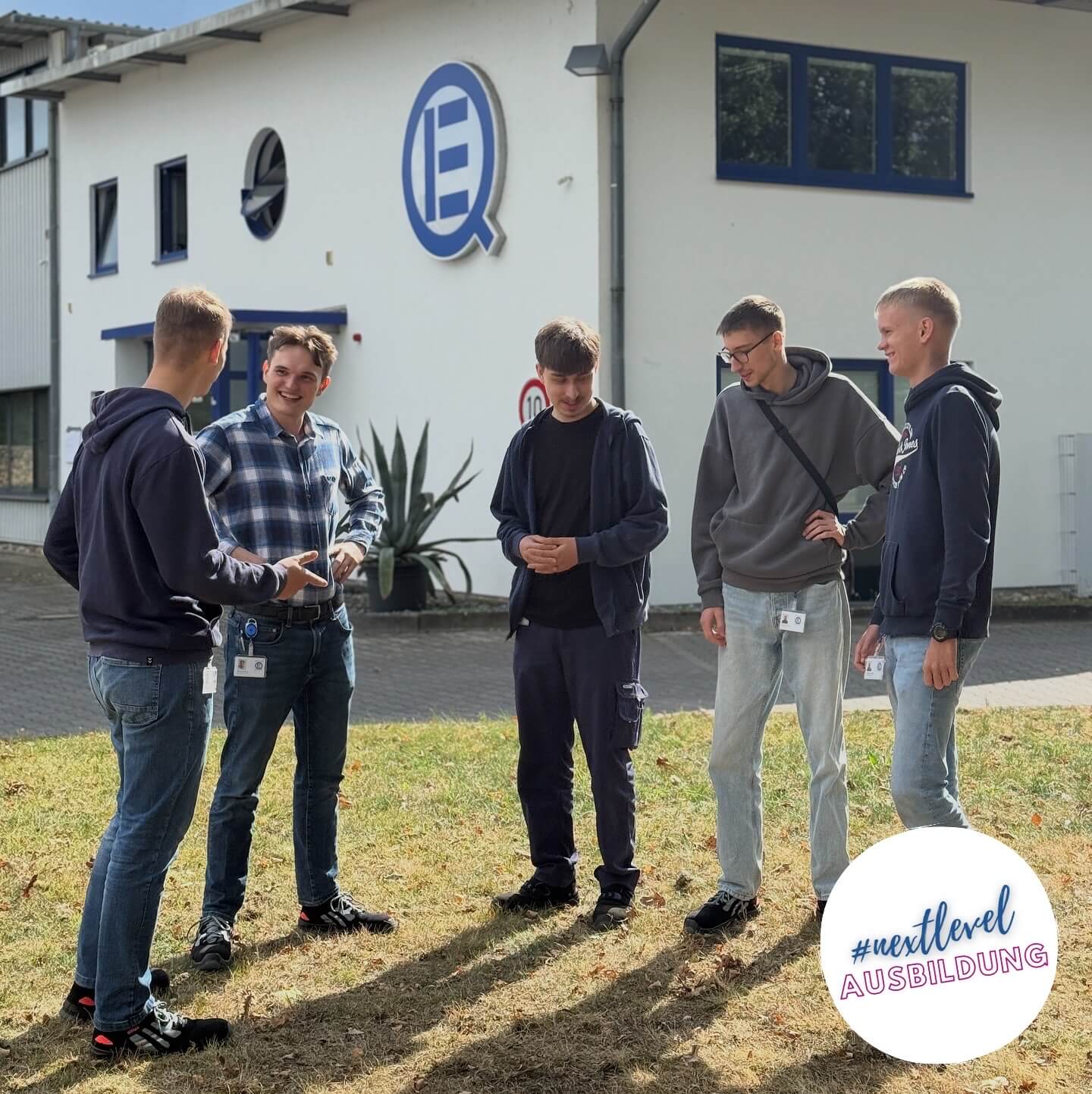 Fuenf junge Maenner mit hellen Hosen und dunklen Pullovern lachen im Hof vor einem Firmengebaeude