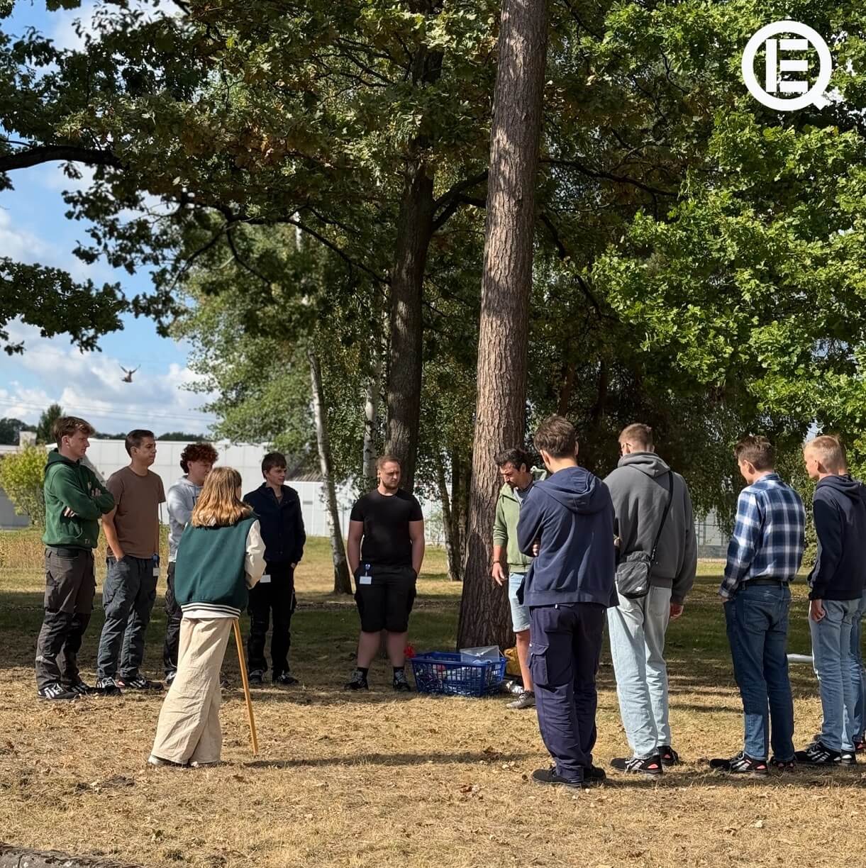 Eine Gruppe junger Menschen steht entspannt im Kreis im Schatten hoher Bäume