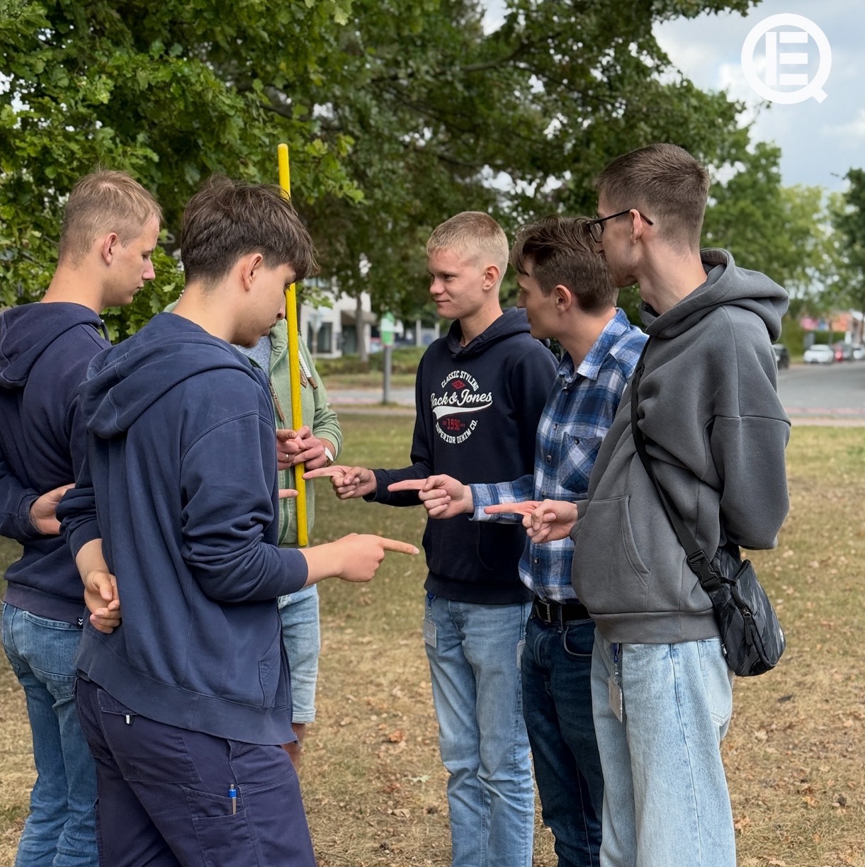Sechs junge Männer besprechen sich, einige tragen blaue Kleidung im Freien.